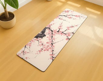 May include: A rectangular yoga mat featuring a floral design with pink blossoms and dark branches against a cream background. The mat has a black border and is placed on a light wood floor. A potted plant is visible in the background.