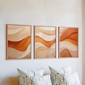 May include: Three framed abstract art prints with warm tones of beige, orange, and brown. The artwork features flowing, wavy patterns. The frames are a light wood color, and the prints are hanging above decorative pillows.