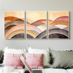 Op de afbeelding: Drie ingelijste abstracte kunstprints. Elke print toont een aquarel landschap van gelaagde, gebogen heuvels in tinten bruin, roze en grijs, tegen een warme gele lucht. Het kunstwerk wordt tentoongesteld boven een witte bank met roze en grijze kussens.