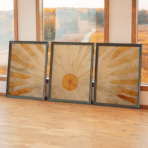 May include: Three framed wall art pieces with a sunburst design. The artwork features a central circular element with radiating lines in shades of yellow and orange. The frames are dark gray, and the pieces are leaning against a window.