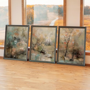 May include: Three framed abstract landscape paintings. The artwork features muted tones of green, brown, and peach, with a focus on natural elements like trees and water. The frames are dark, and the paintings are leaning against a wall.