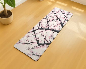 May include: A rectangular yoga mat featuring a watercolor design of cherry blossom branches with pink flowers and black branches against a white background. The mat is on a light wood floor.