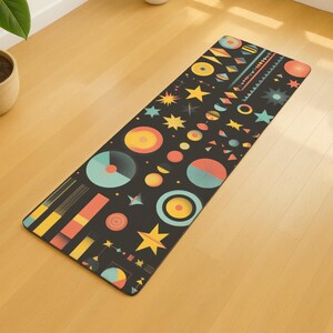May include: A rectangular yoga mat with a black background and a colorful geometric pattern. The design includes circles, stars, and other shapes in shades of yellow, orange, teal, and red. The mat is lying on a light-colored wooden floor.