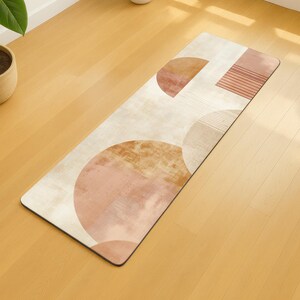 May include: Rectangular yoga mat featuring an abstract design with peach, brown, and tan geometric shapes on a cream background. The design includes circles and lines. The mat is on a wooden floor. Dimensions are approximately 173 cm long.