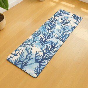 May include: A rectangular yoga mat with a blue coral reef design. The pattern features stylized blue coral branches against a white and light blue watercolor background. The mat is on a light wood floor.