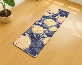 May include: A rectangular yoga mat with a floral design. The mat features large yellow and peach-colored chrysanthemum flowers and green leaves on a dark blue background. The mat is placed on a light-colored wooden floor.