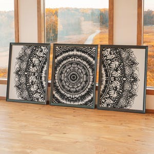May include: Three framed art prints with intricate black and white mandala designs. The artwork is displayed on a light-colored background, framed in dark gray, and set against a backdrop of a window overlooking a landscape.