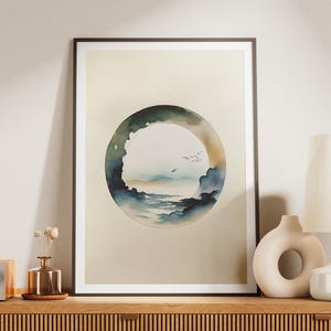 May include: Framed watercolor art print featuring a circular landscape scene with a mountain range, clouds, and birds. The artwork is in shades of blue, green, and beige, and is displayed on a wooden surface.