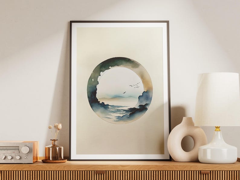 May include: Framed watercolor art print featuring a circular landscape scene with a mountain range, clouds, and birds. The artwork is in shades of blue, green, and beige, and is displayed on a wooden surface.