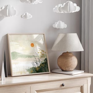 May include: A framed watercolor painting of a landscape with a large orange sun, green hills, and a body of water. The painting is hanging on a light wood wall with a lamp and a book on a table below it.