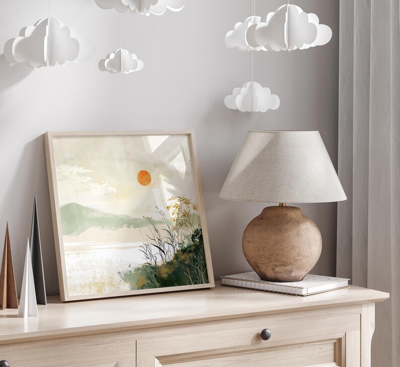 May include: A framed watercolor painting of a landscape with a large orange sun, green hills, and a body of water. The painting is hanging on a light wood wall with a lamp and a book on a table below it.