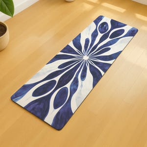 Puede incluir: Una esterilla de yoga rectangular con un diseño floral azul y blanco. El patrón irradia desde un punto central, con elementos alargados en forma de lágrima en tonos azules sobre un fondo blanco. La esterilla está sobre un suelo de madera clara.