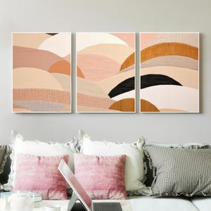 May include: Three framed abstract art prints. The artwork features a landscape design with layered shapes in shades of peach, tan, and black. The prints are displayed above a sofa with pink and gray pillows.