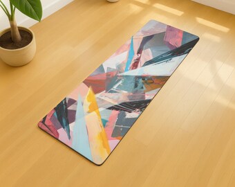May include: Rectangular yoga mat with an abstract geometric design. The mat features a colorful pattern of overlapping shapes in shades of pink, blue, yellow, and black. The mat is placed on a wooden floor.