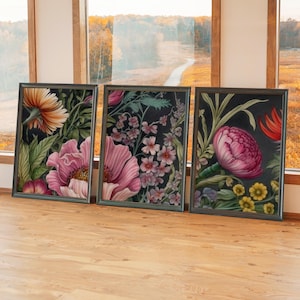 May include: Three framed floral art prints. Each features a dark background with colorful flowers in shades of pink, orange, and yellow. The frames are dark, and the prints are displayed on a wooden surface near a window.