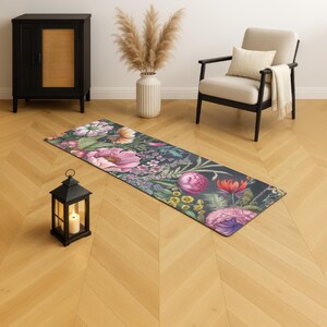 May include: A floral yoga mat with a dark background and a vibrant array of pink, purple, and yellow flowers. The mat is rectangular and lies on a wooden floor. A black lantern with a lit candle sits nearby.