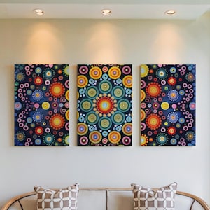 May include: Three rectangular canvas artworks featuring vibrant, circular patterns in blue, orange, yellow, and pink. The paintings are mounted on a white wall above a sofa with patterned cushions. Recessed lighting illuminates the art.