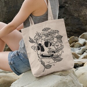May include: A beige tote bag with a black and white illustration of a skull with mushrooms growing out of it. The bag is sitting on a rock by the ocean.