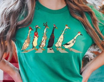 Peut inclure: Un t-shirt vert avec un motif de Noël représentant un groupe de canards portant des bonnets de Père Noël et des lumières de Noël traversant un passage pour piétons.