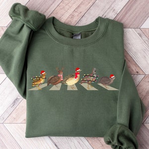 May include: An olive green crewneck sweatshirt featuring a graphic print of five quails walking in a line across a crosswalk. The birds are wearing festive accessories including red Santa hats, a red and white striped scarf, and colorful string lights.