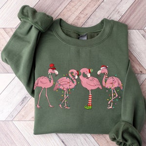 May include: A green sweatshirt with a Christmas-themed design featuring four pink flamingos wearing Santa hats and adorned with Christmas lights.