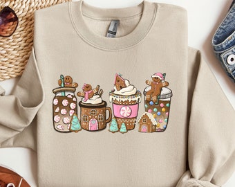 May include: A beige sweatshirt with a festive design featuring gingerbread men, houses, and mugs of hot chocolate topped with whipped cream and candy.