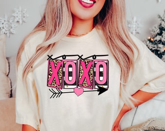 May include: A white t-shirt with a pink and black "XOXO" graphic featuring an arrow and heart. The graphic has a leopard print design.