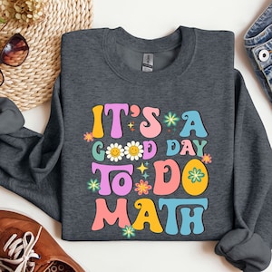 May include: A dark gray sweatshirt with the text "It's a good day to do math" in colorful retro-style lettering. The text is surrounded by flowers and stars.