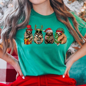 May include: A green t-shirt with a graphic of five sloths wearing Christmas hats and holding Christmas decorations. The sloths are wearing a Santa hat, a reindeer headband, a top hat, a green elf hat, and sunglasses.