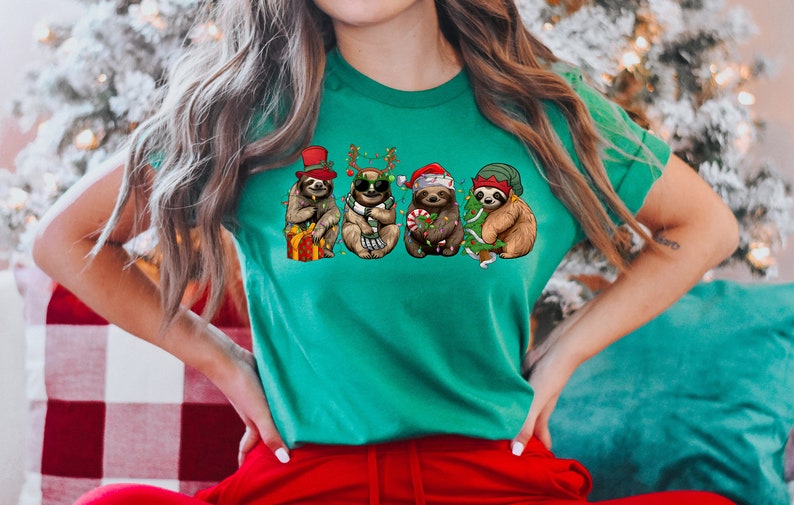 May include: A green t-shirt with a graphic of five sloths wearing Christmas hats and holding Christmas decorations. The sloths are wearing a Santa hat, a reindeer headband, a top hat, a green elf hat, and sunglasses.