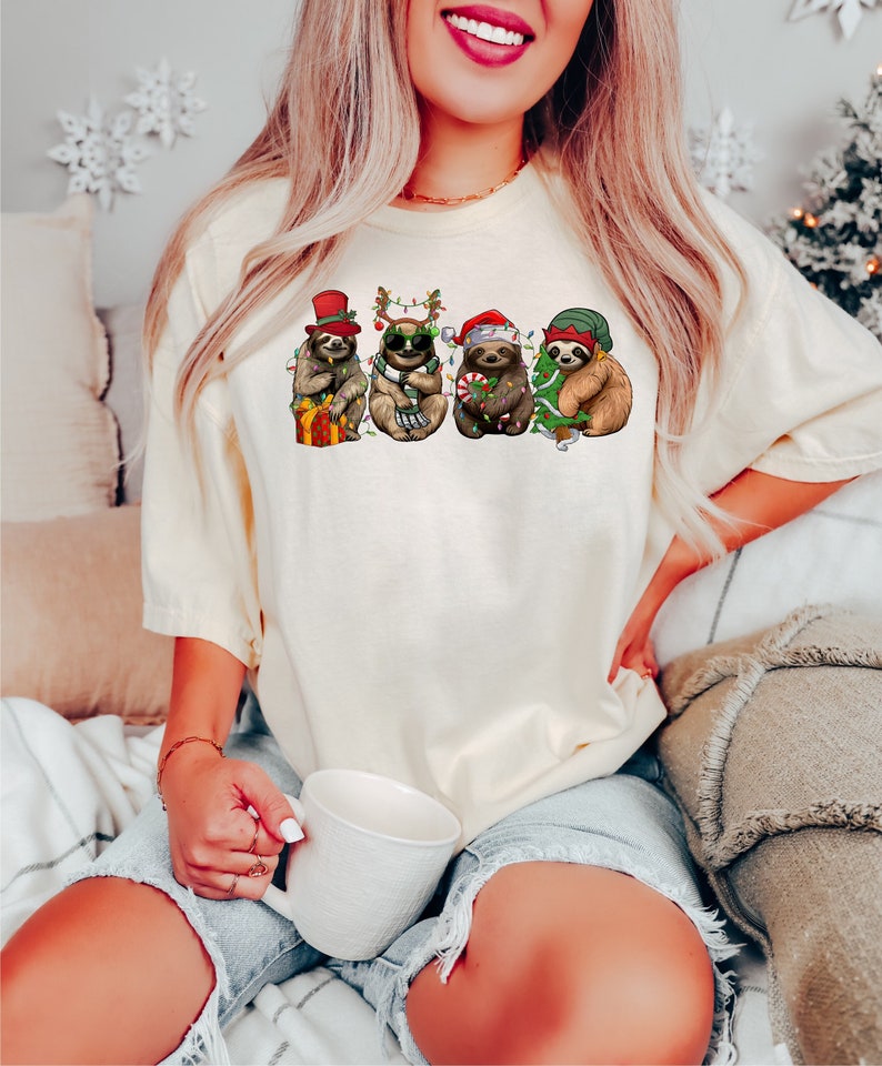 May include: A white t-shirt with a graphic of four sloths wearing Christmas hats and decorations. The sloths are holding gifts, candy canes, and a Christmas tree.