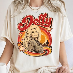 May include: A cream-colored short-sleeve t-shirt featuring a graphic print of a woman with voluminous blonde hair wearing a western-style shirt. The design includes the word Dolly in red, surrounded by orange and yellow sunset stripes and stars.