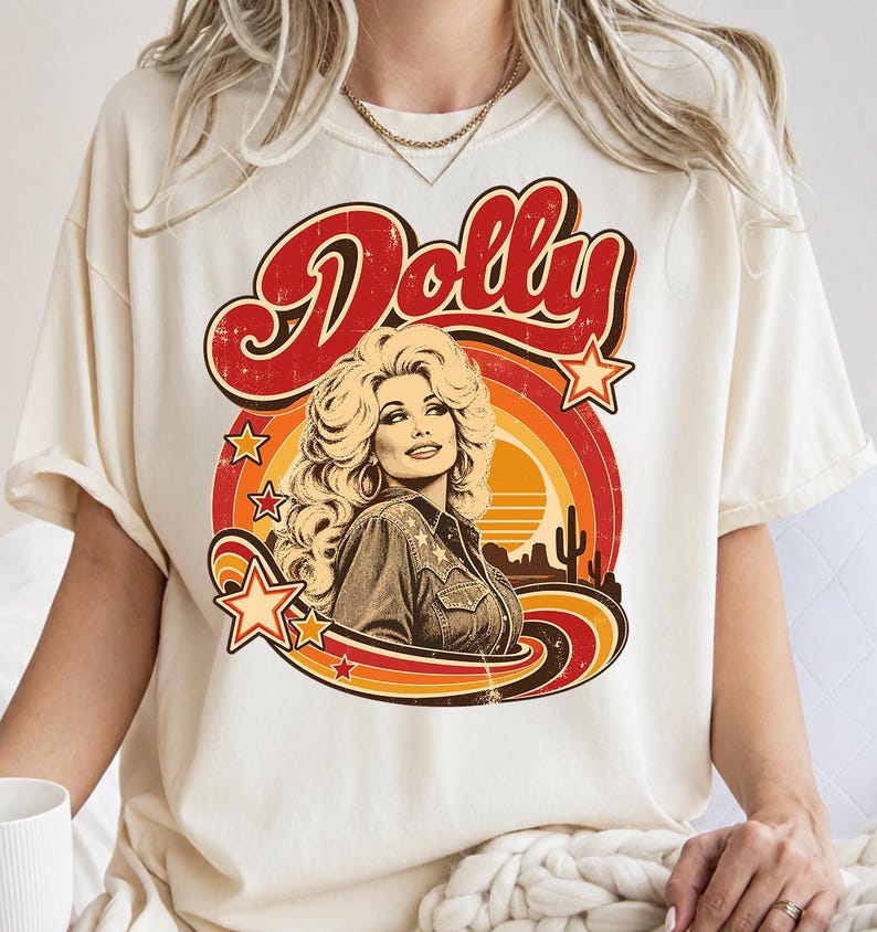 May include: A cream-colored short-sleeve t-shirt featuring a graphic print of a woman with voluminous blonde hair wearing a western-style shirt. The design includes the word Dolly in red, surrounded by orange and yellow sunset stripes and stars.