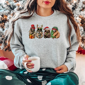 May include: A gray sweatshirt with a Christmas-themed design featuring four sloths wearing festive hats and holding gifts. The sloths are surrounded by Christmas lights and greenery.
