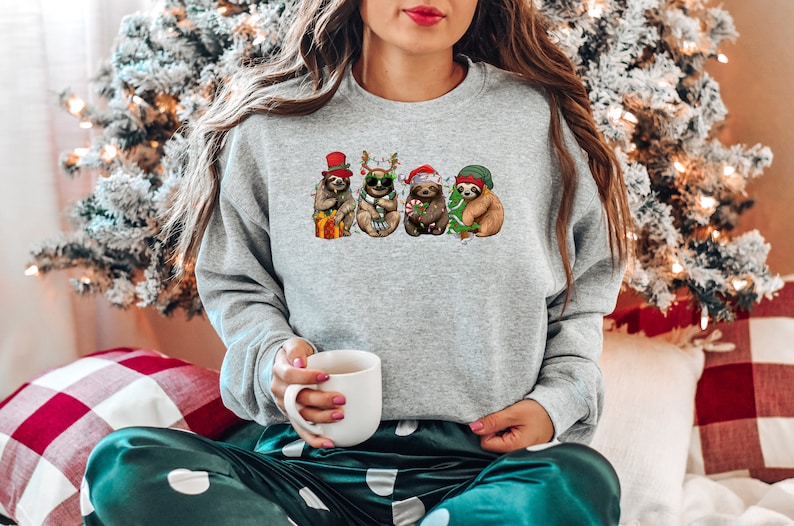May include: A gray sweatshirt with a Christmas-themed design featuring four sloths wearing festive hats and holding gifts. The sloths are surrounded by Christmas lights and greenery.