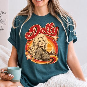 May include: A dark teal short-sleeve t-shirt featuring a graphic print of a woman with blonde hair and the text Dolly. The design includes a circular orange and yellow sunset background with stars, a desert landscape, and a cactus silhouette.