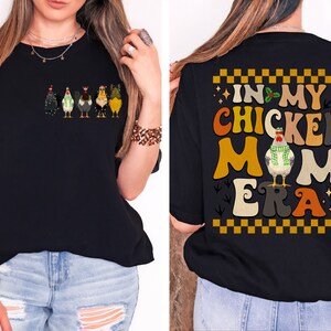 May include: Black t-shirt with a graphic of a line of chickens wearing Christmas lights and hats on the front. The back of the shirt has a graphic of a chicken wearing a scarf and the text "In My Chicken Mom Era" in a retro font.