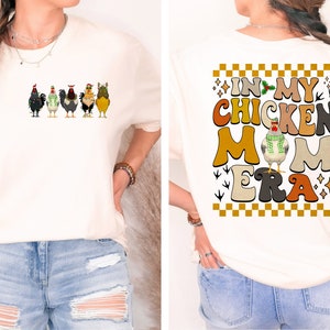 May include: A white t-shirt with a graphic of chickens and the text "In My Chicken Mom Era" on the back. The graphic is in a retro style with a checkered background.