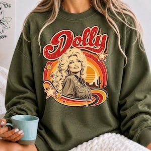 May include: An olive green crewneck sweatshirt featuring a graphic print of Dolly Parton with the text "Dolly" in red script. The design includes a retro sunset, stars, and a desert landscape with a cactus in shades of orange, yellow, and red.