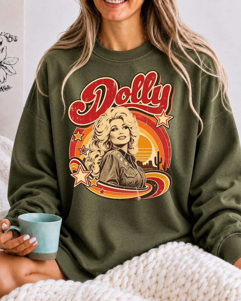 May include: An olive green crewneck sweatshirt featuring a graphic print of Dolly Parton with the text "Dolly" in red script. The design includes a retro sunset, stars, and a desert landscape with a cactus in shades of orange, yellow, and red.