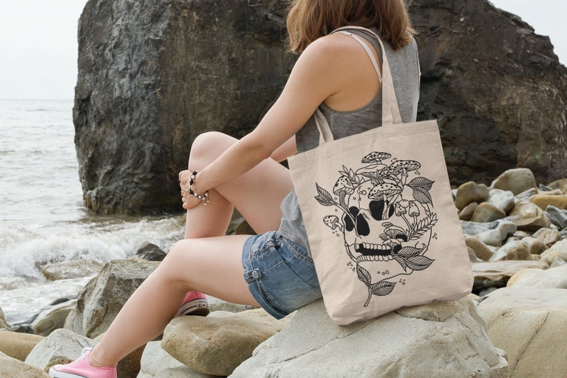 May include: A beige tote bag with a black and white illustration of a skull with mushrooms growing out of it. The bag is sitting on a rock by the ocean.