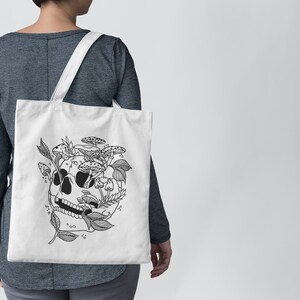 May include: A white tote bag with a black and white illustration of a skull with mushrooms and leaves growing out of it.