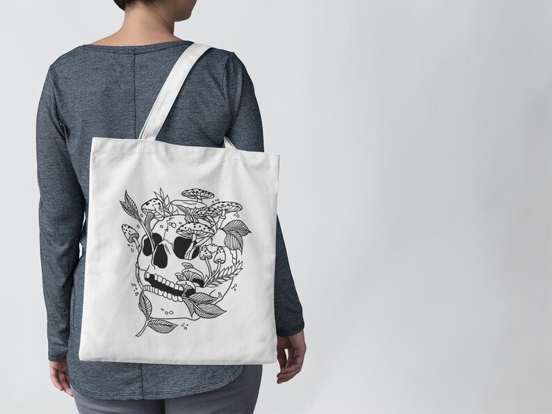 May include: A white tote bag with a black and white illustration of a skull with mushrooms and leaves growing out of it.