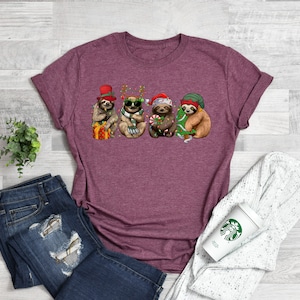 May include: A maroon t-shirt with a graphic of four sloths wearing Christmas hats and holding Christmas decorations. The sloths are wearing Santa hats, reindeer antlers, a top hat, and a green hat.