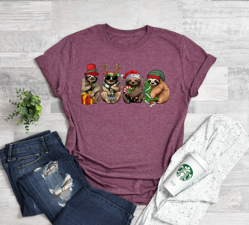 May include: A maroon t-shirt with a graphic of four sloths wearing Christmas hats and holding Christmas decorations. The sloths are wearing Santa hats, reindeer antlers, a top hat, and a green hat.