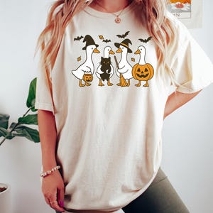 Pu&ograve; includere: T-shirt beige con grafica a tema Halloween. Il design include quattro anatre dei cartoni animati vestite con cappelli da strega, che tengono in mano una zucca, un gatto nero e una scopa. Sono inclusi anche pipistrelli e foglie.