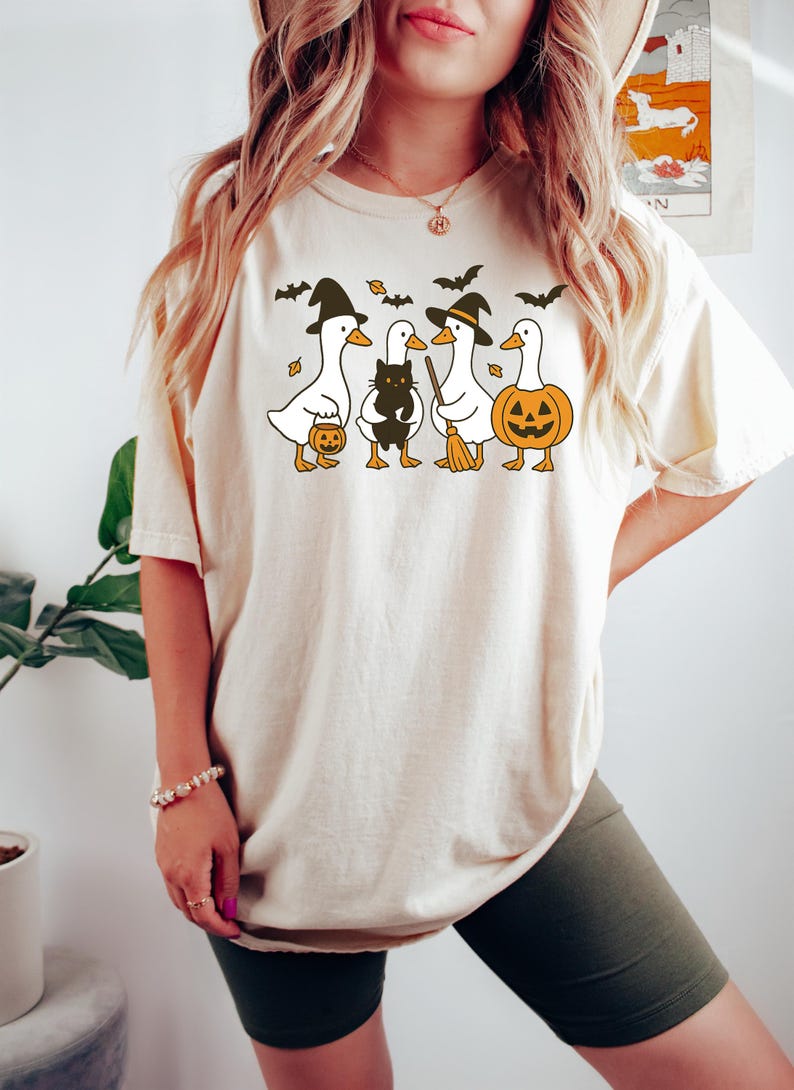 Pu&ograve; includere: T-shirt beige con grafica a tema Halloween. Il design include quattro anatre dei cartoni animati vestite con cappelli da strega, che tengono in mano una zucca, un gatto nero e una scopa. Sono inclusi anche pipistrelli e foglie.