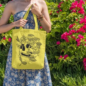 May include: A yellow tote bag with a black and white illustration of a skull with mushrooms growing out of it. The bag has a floral pattern and is being held by a person.