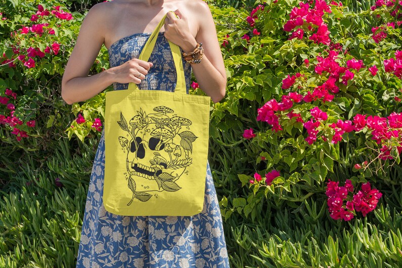 May include: A yellow tote bag with a black and white illustration of a skull with mushrooms growing out of it. The bag has a floral pattern and is being held by a person.