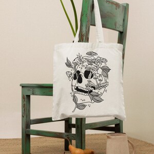 May include: White tote bag with a black line drawing of a skull with mushrooms growing out of it. The bag is sitting on a green chair with a white flower in the background.